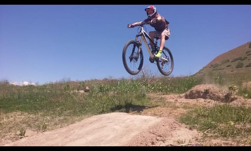 A person in a helmet jumps off a dirt ramp on a mountain bike, with both wheels off the ground against a clear blue sky and grassy background. The DT-DH mountain bike trail.