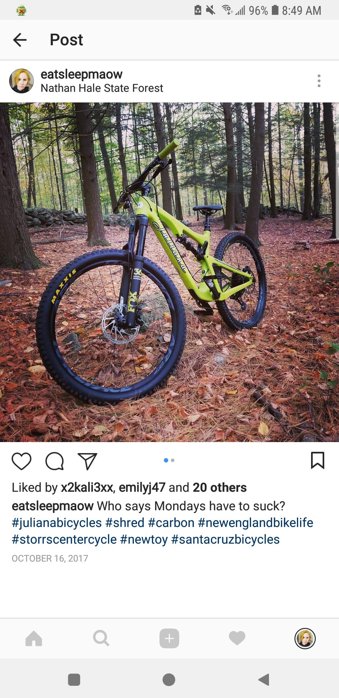 Juliana Roubion: A bright green mountain bike is positioned in a wooded area with tall trees and fallen leaves. The bike features thick tires and a front suspension fork. In the background, there are stone outcroppings and more trees, creating a natural setting.