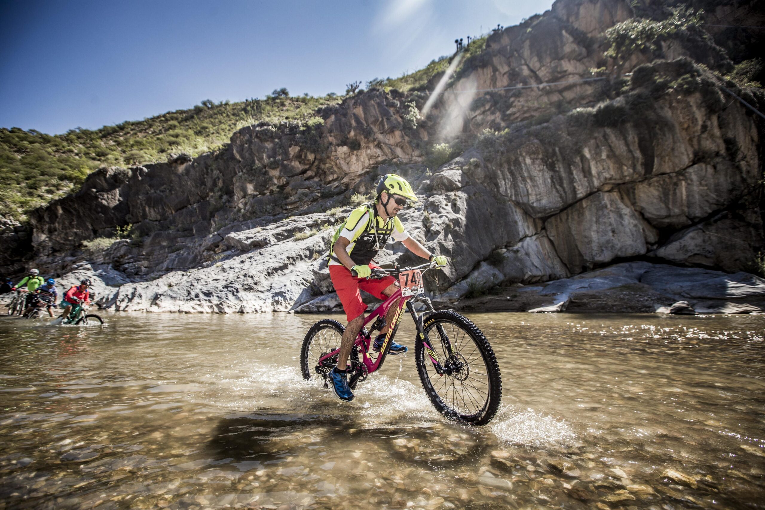 Santa Cruz Bronson C: A mountain biker riding through shallow water on a rocky trail, surrounded by tall cliffs and greenery under a clear blue sky. The cyclist, wearing a helmet and bright protective gear, splashes the water as they navigate the terrain. Other bikers can be seen in the background.