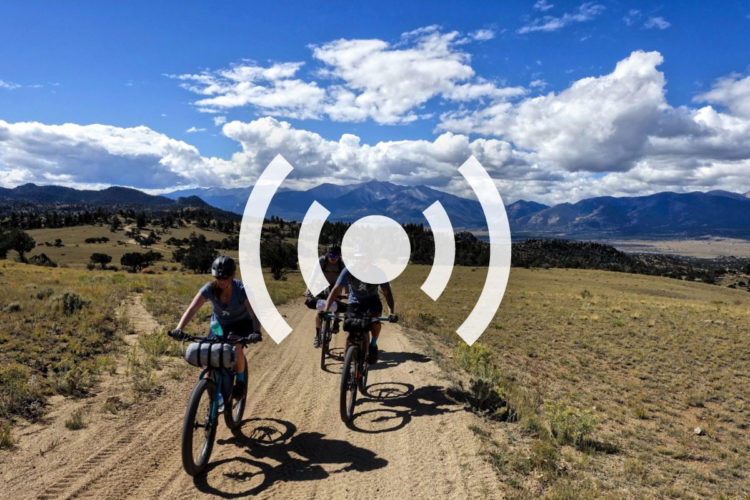 Two cyclists ride on a dirt path through a scenic landscape featuring rolling hills and distant mountains. The sky is bright blue with fluffy white clouds scattered throughout. The cyclists are equipped with helmets and bags on their bikes, enjoying a sunny outdoor adventure.
