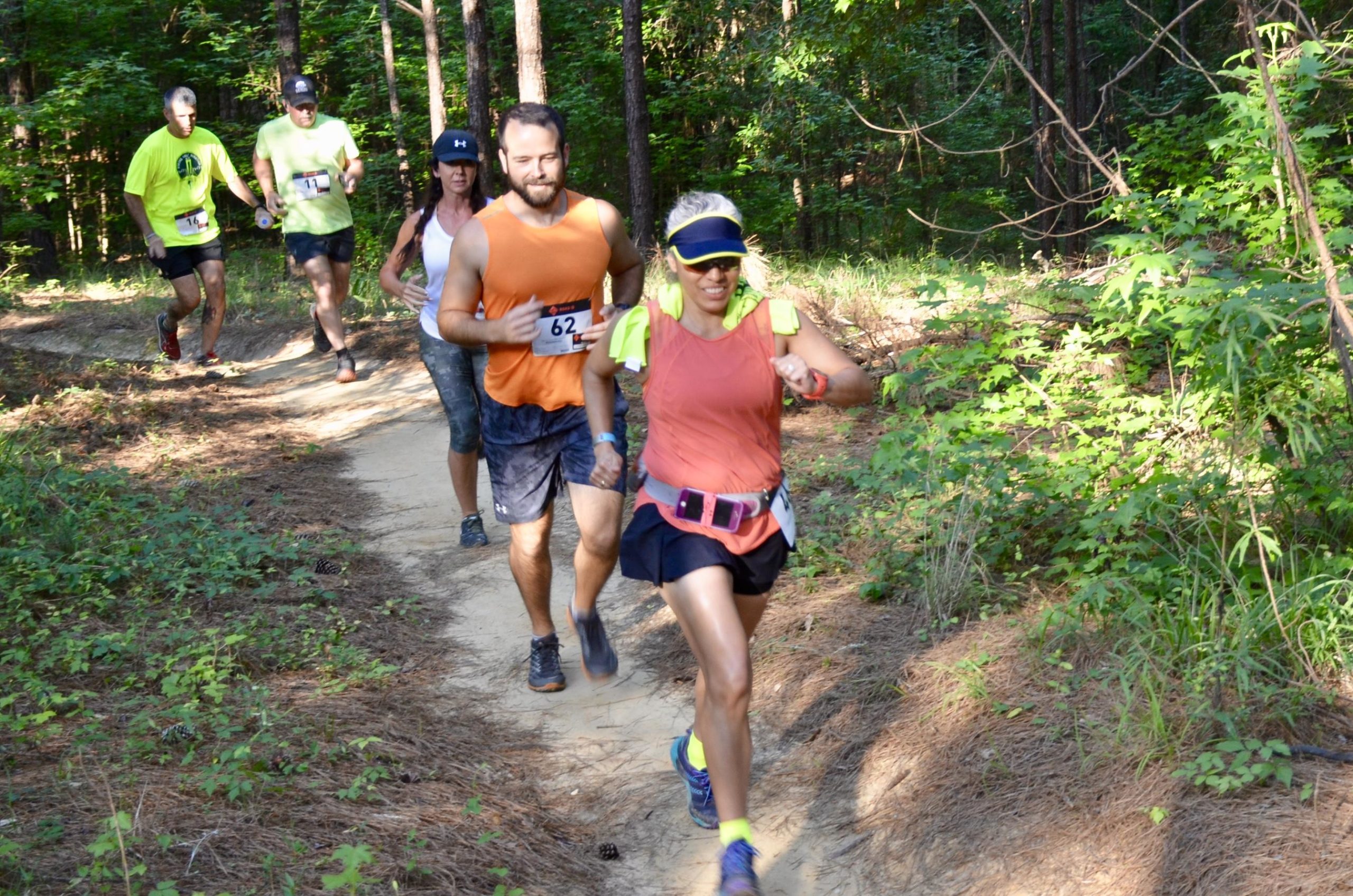 A group of runners participating in a race on a dirt trail surrounded by trees. The runners are wearing athletic gear, including tank tops and shorts, and are focused on the path ahead. The scene is set in a wooded area, with greenery and pine needles lining the trail. Mt. Zion Bike Trails mountain bike trail.