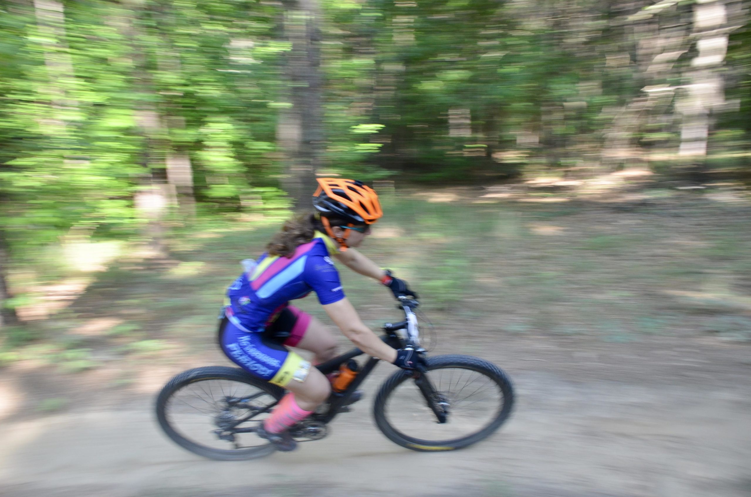 A cyclist in motion rides a mountain bike along a dirt path surrounded by greenery. The athlete is wearing a colorful cycling jersey and protective gear, including an orange helmet. The image conveys a sense of speed and adventure in a natural setting. Mt. Zion Bike Trails mountain bike trail.