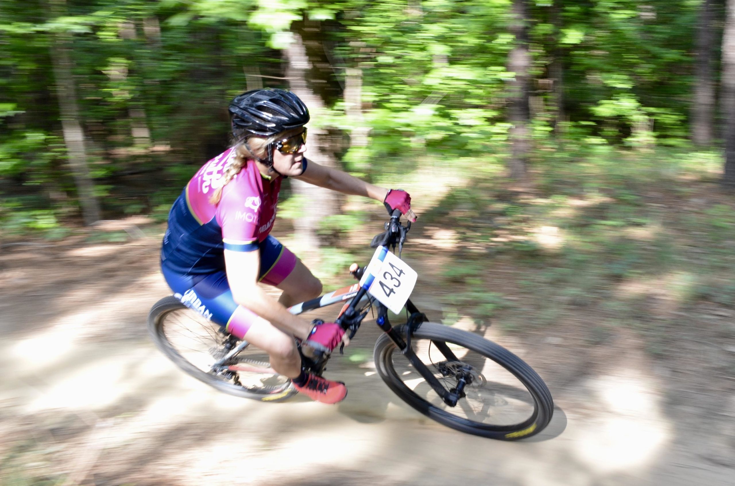 A cyclist in a bright pink and blue jersey speeds along a dirt trail surrounded by green trees. The image captures the motion blur, emphasizing the cyclist's fast pace as they maneuver the terrain. The bike has a race number displayed on the front. Mt. Zion Bike Trails mountain bike trail.
