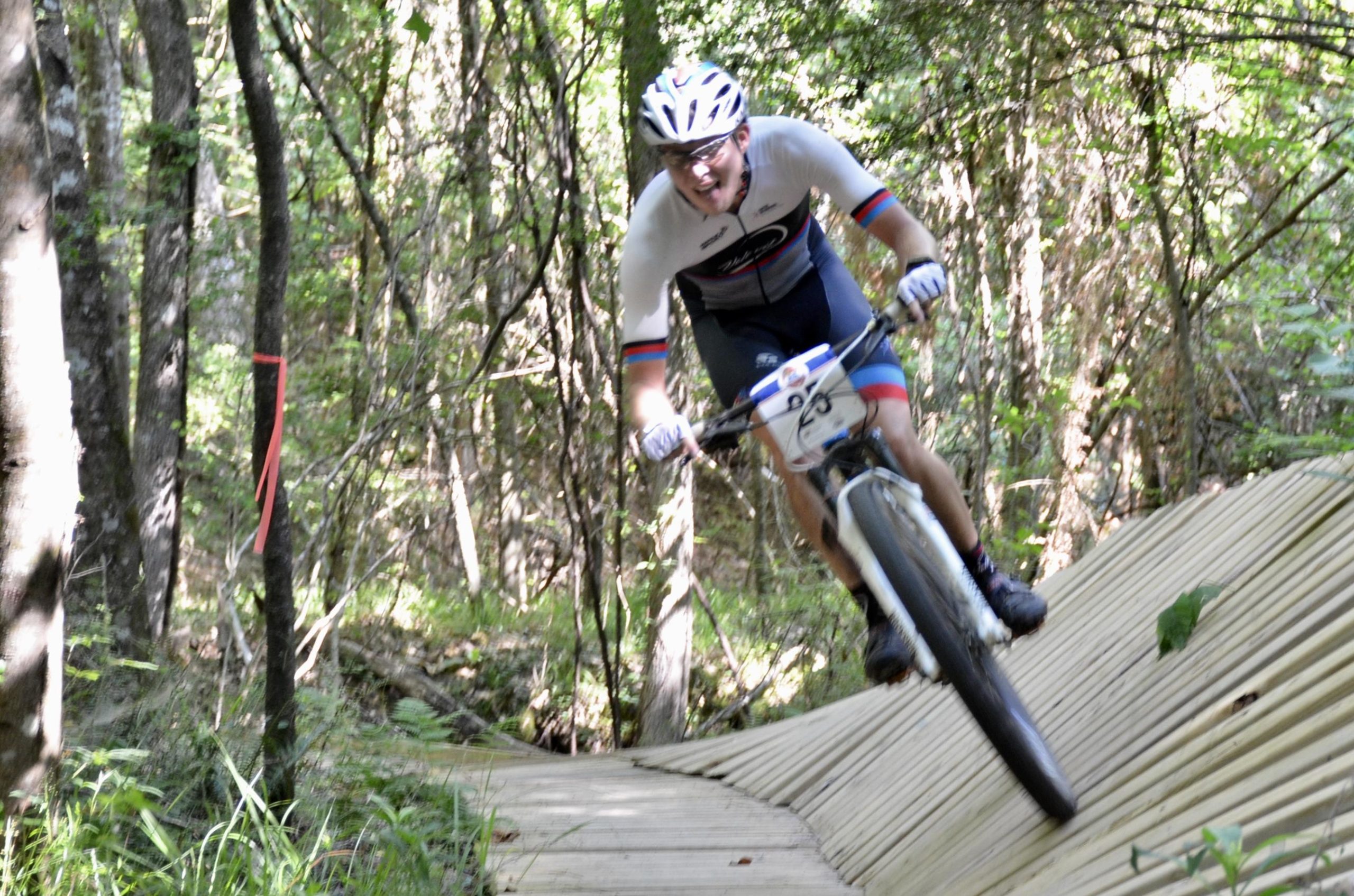 A mountain biker rides energetically on a wooden trail through a forest. The rider is wearing a white jersey with colorful stripes and a helmet, showing intense concentration and joy as he navigates the winding path. Surrounding trees and greenery create a vibrant natural backdrop. Mt. Zion Bike Trails mountain bike trail.