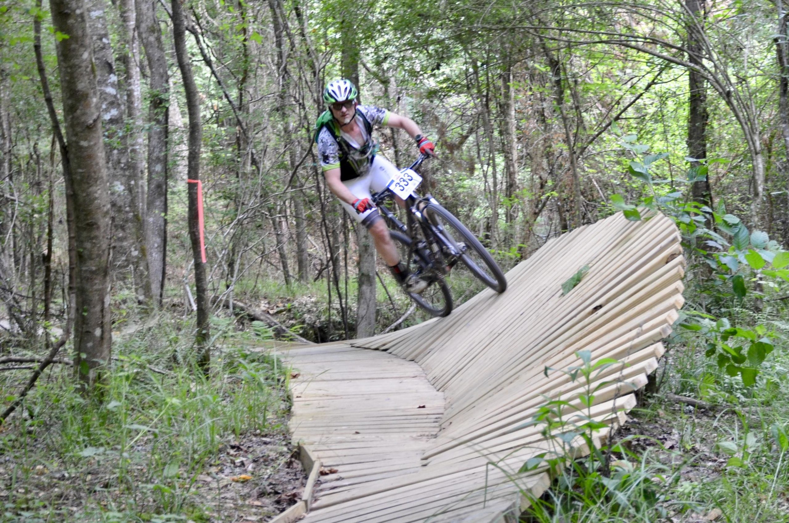 A mountain biker navigating a wooden bike trail feature in a forested area, with trees and greenery surrounding the path. The cyclist is wearing a helmet and cycling gear, appearing to maneuver the curved wooden structure with skill and speed. A pink marker is visible alongside the trail. Mt. Zion Bike Trails mountain bike trail.