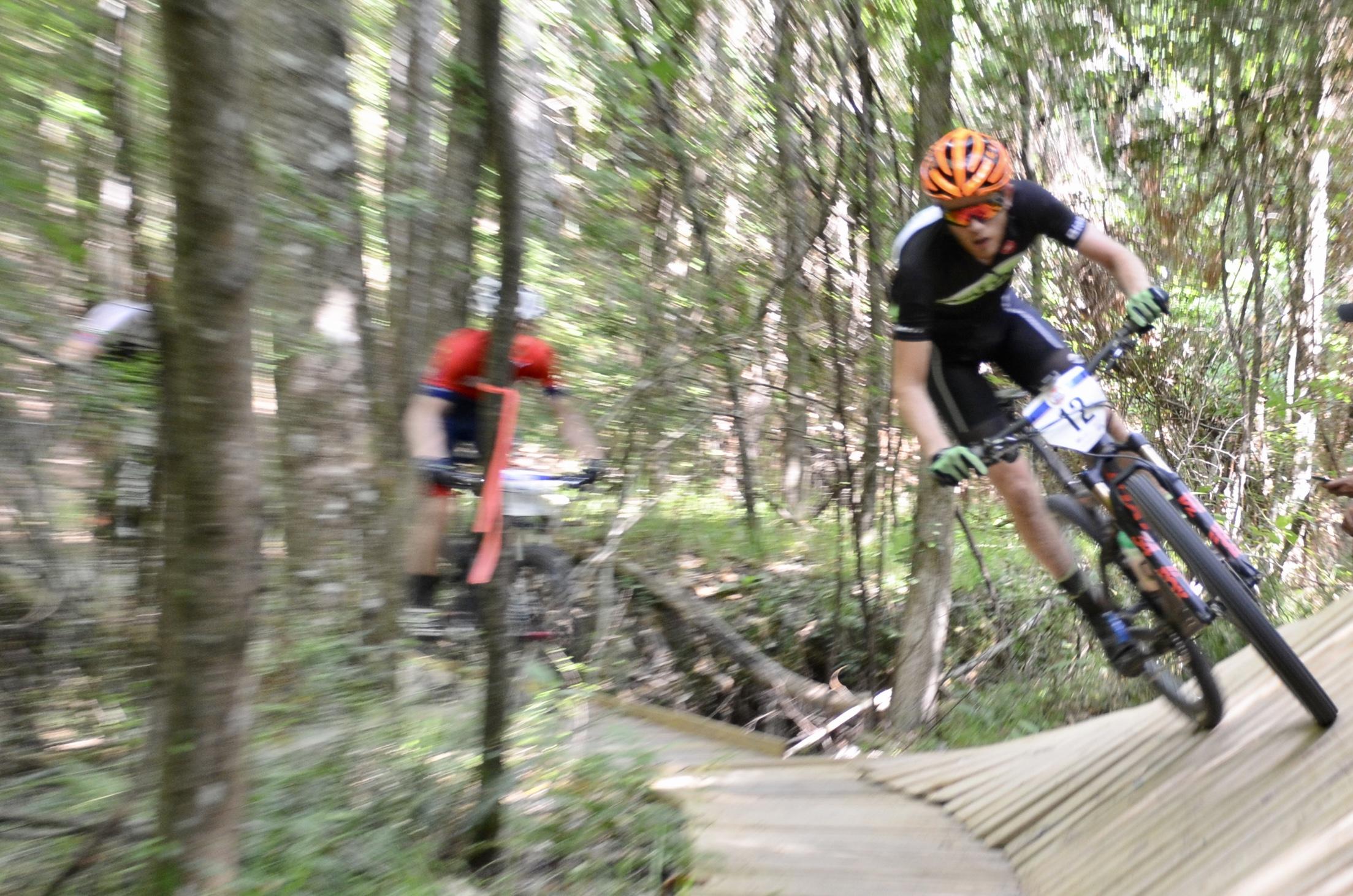 A mountain biker in a black jersey and orange helmet navigates a wooden ramp on a trail surrounded by trees, demonstrating speed and agility. Another cyclist in a red jersey can be seen blurred in the background, indicating the fast-paced nature of the race. Mt. Zion Bike Trails mountain bike trail.