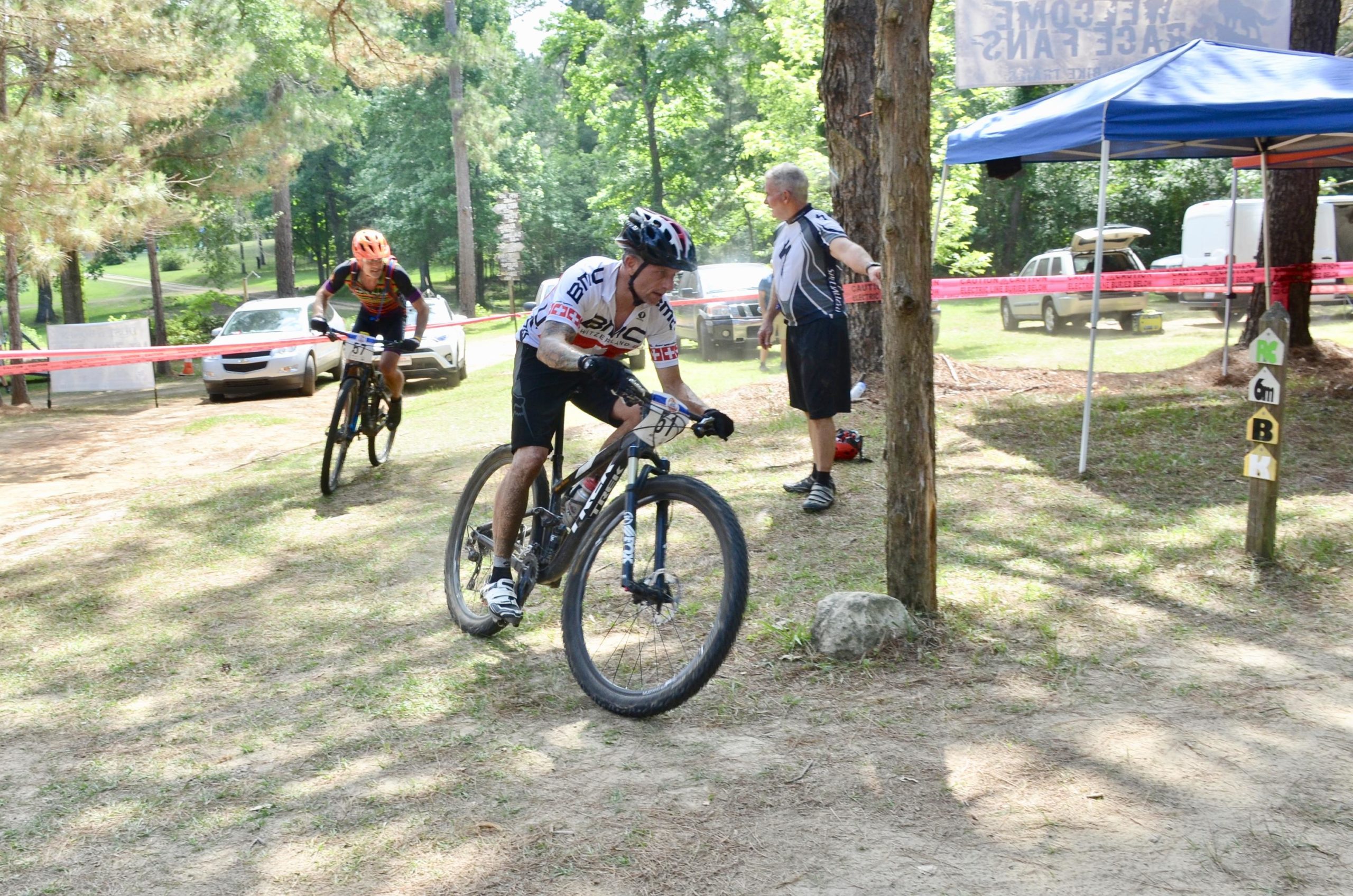 Alt text: Two mountain bikers race through a wooded area during a cycling event, with one cyclist leaning into a turn and another approaching in the background. A spectator stands near a blue tent, and trail markers are visible on the ground. Mt. Zion Bike Trails mountain bike trail.