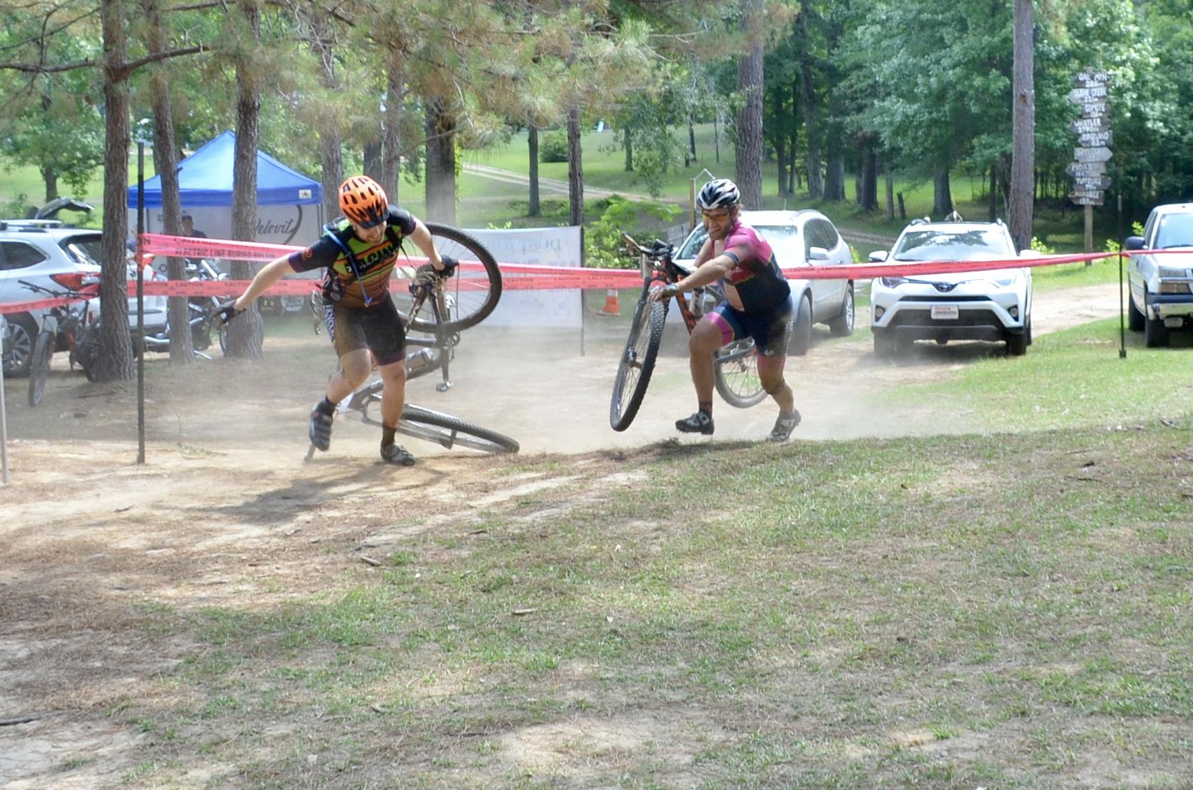 Two mountain bikers are participating in a race, one of whom has fallen off their bike and is lifting it in a cloud of dust while the other is navigating past. The scene takes place on a grassy track with trees and parked cars in the background, and there are red barriers marking the race route. Mt. Zion Bike Trails mountain bike trail.