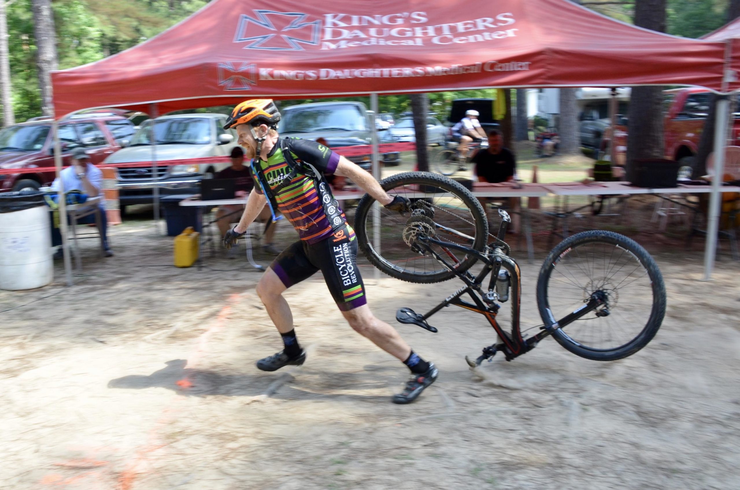 A cyclist in a colorful jersey runs while carrying a bicycle wheel, with a tent advertising "King's Daughters Medical Center" in the background. The scene is set outdoors, with parked vehicles and spectators in the background. The cyclist appears to be participating in a race or event, showcasing a dynamic and energetic moment. Mt. Zion Bike Trails mountain bike trail.