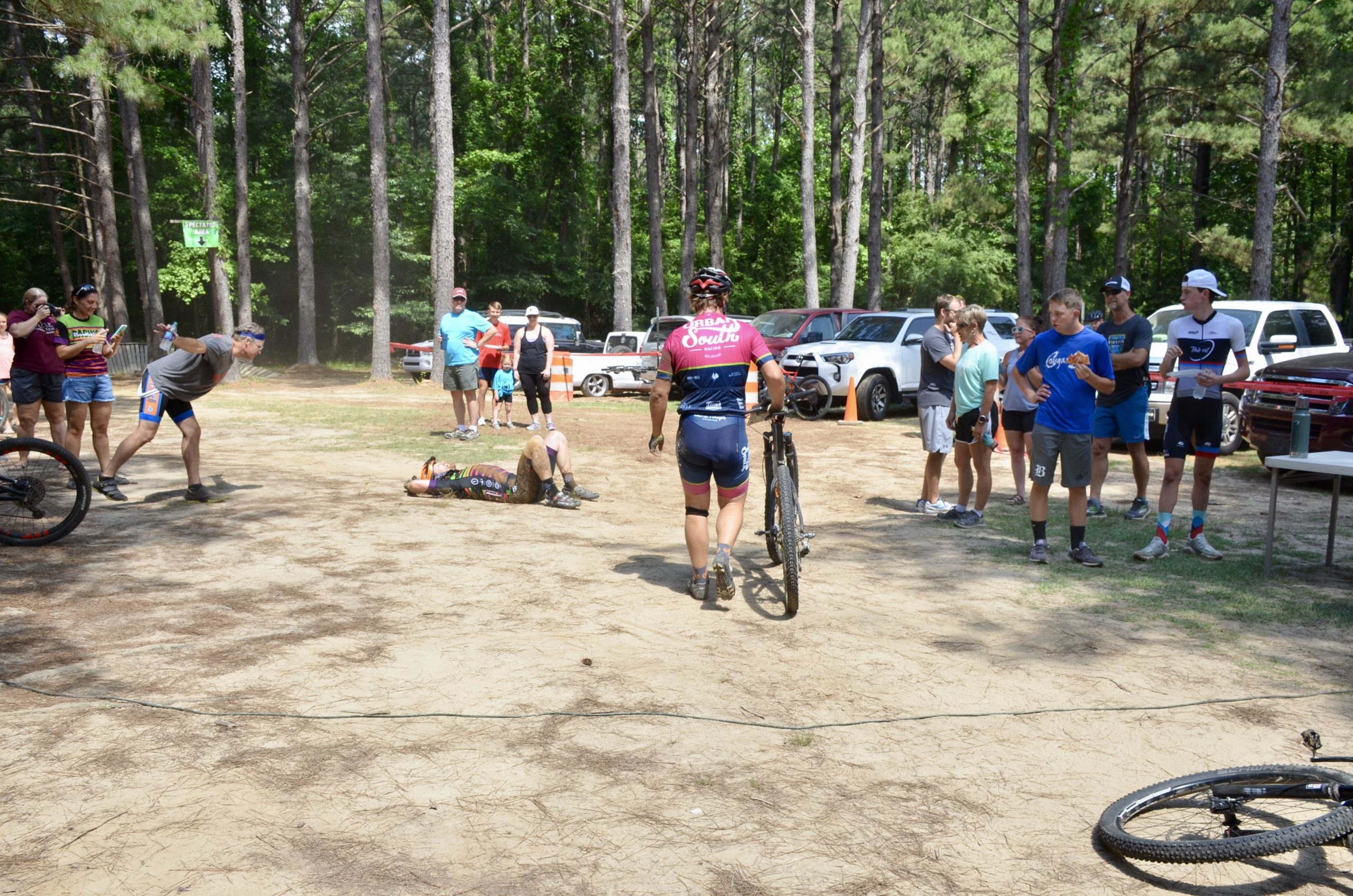 A group of people gathered in a forested area, watching a cycling event. In the foreground, a cyclist is lying on the ground, while another cyclist walks away with a bike. Several spectators, including men and women of various ages, are engaged with the scene, some holding signs. Vehicles are parked in the background, and the atmosphere appears lively and focused on the sporting event. Mt. Zion Bike Trails mountain bike trail.