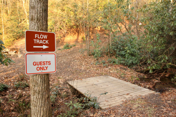 A wooden bridge leading over a small creek, accompanied by two signs on a nearby tree. The top sign indicates "FLOW TRACK" with an arrow pointing right, while the bottom sign reads "GUESTS ONLY." The surrounding area features leaf-covered ground and lush foliage, suggesting a natural outdoor setting.