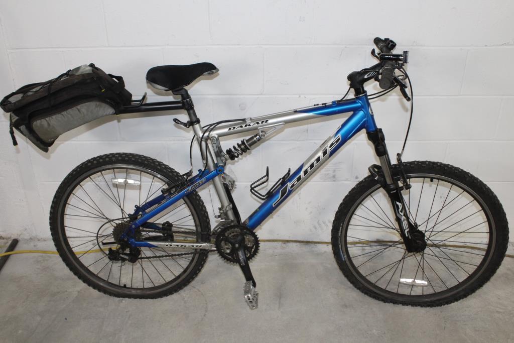 A blue and silver mountain bike with a rear saddlebag, positioned on a concrete floor against a plain white wall. The bike features thick, knobby tires, dual shock absorbers, and a gear system. Cedarville State Forest mountain bike trail.
