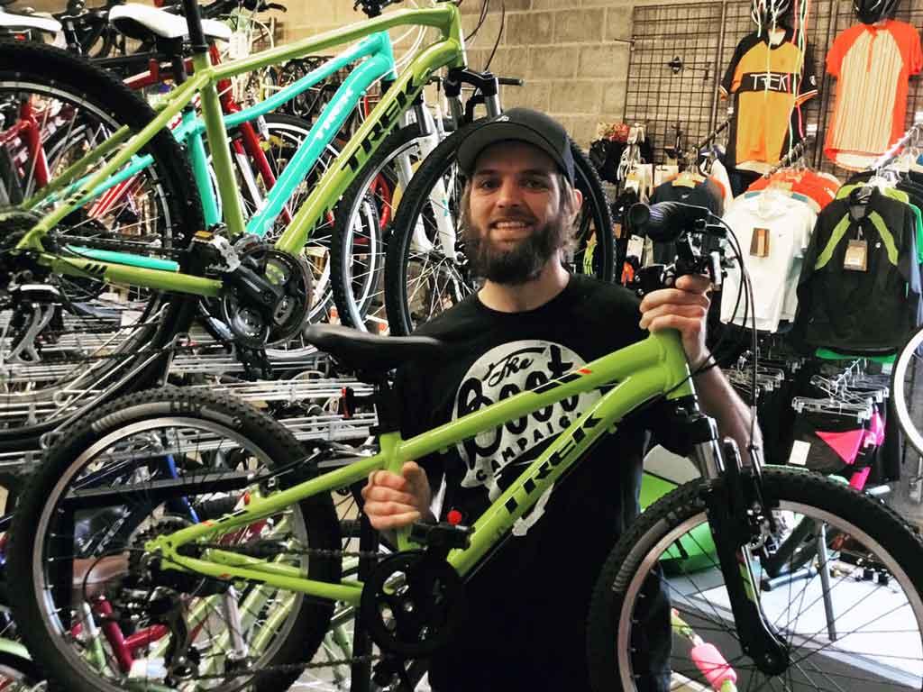A person with a beard and a cap smiles while holding a green bicycle frame in a bike shop, surrounded by various bicycles and cycling apparel.