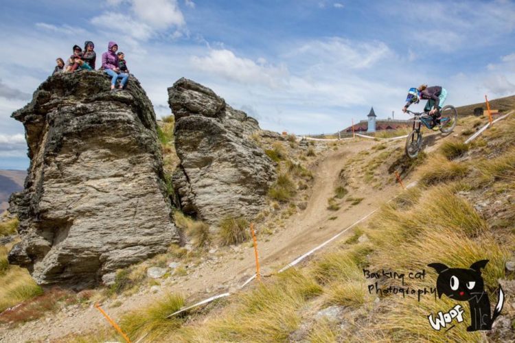 A mountain biker jumps off a rocky ledge while descending a dirt track. In the background, a group of spectators sits atop a large rock, watching the action. The scene is set against a backdrop of rolling hills and a cloudy sky. Orange markers line the track, indicating the course.