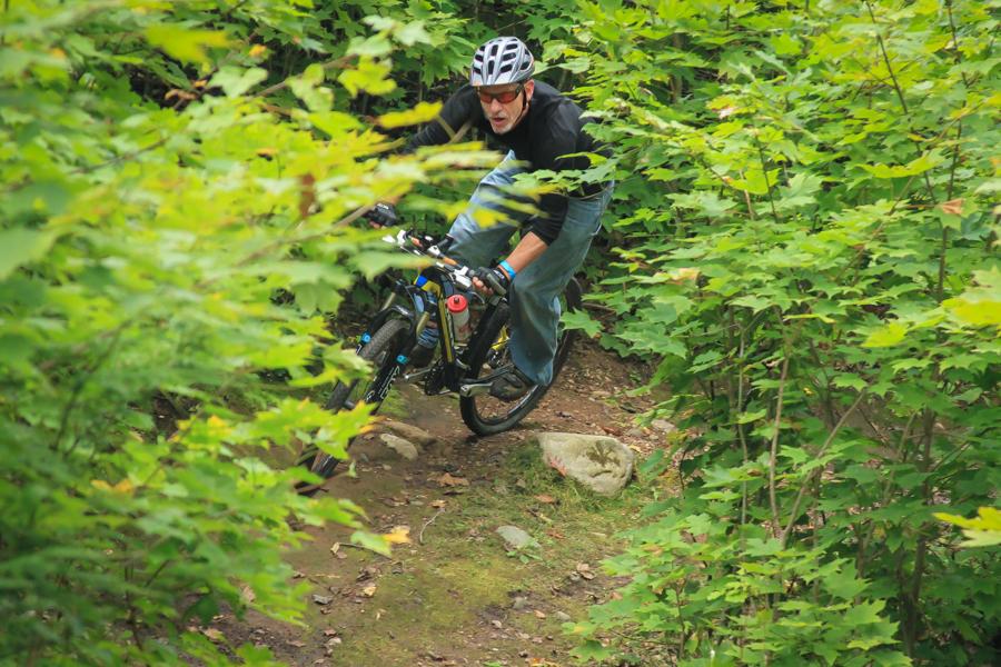 A mountain biker navigating a narrow trail surrounded by dense green foliage, leaning into a turn while riding over rocky terrain. The cyclist is wearing a helmet and has an intense focus on the path ahead. Raven Trails mountain bike trail.