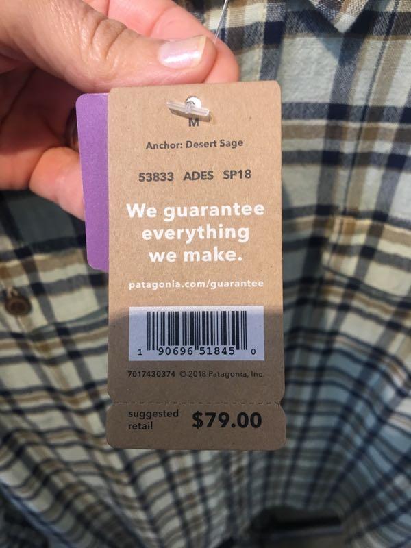 A close-up view of a clothing tag being held in a hand, featuring information on a Patagonia item. The tag indicates the size (M), color (Desert Sage), product number, and features a guarantee statement reading "We guarantee everything we make." The suggested retail price is listed as $79.00, and a barcode is visible on the tag. The background shows a plaid shirt fabric.