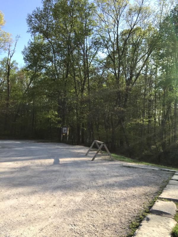 A gravel parking area bordered by trees under clear blue skies, with a wooden barrier in the foreground and a sign partially visible on the left. The scene is bright and sunny, suggesting a peaceful outdoor environment. Brown County Park mountain bike trail.