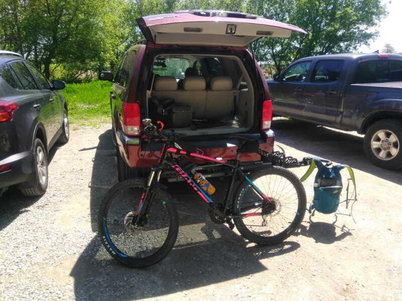 A red SUV with its rear hatch open, revealing a spacious interior, parked in a gravel area. Next to the vehicle, a mountain bike is positioned upright, showcasing its colorful frame and sturdy tires. A backpack is placed on the ground nearby, indicating preparations for an outdoor adventure in a lush, green environment. The Pines mountain bike trail.