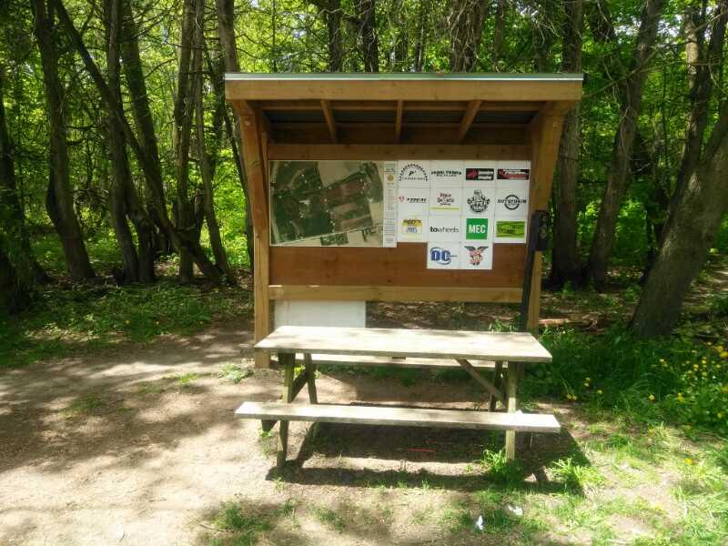 A wooden information kiosk with a thatched roof, featuring a map and various logos affixed to its side, located along a dirt path in a shaded forest area. A picnic-style bench is positioned below the kiosk, surrounded by greenery and trees. The Pines mountain bike trail.