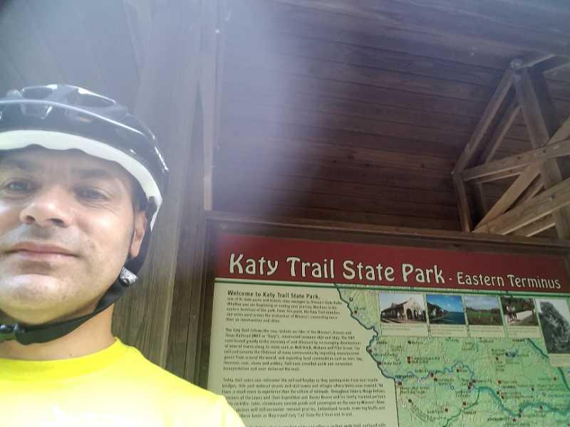 A person wearing a bike helmet and a yellow shirt stands in front of a sign for Katy Trail State Park, Eastern Terminus. The sign provides information about the park and includes a map of the trail. KATY trail mountain bike trail.