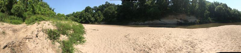 A panoramic view of a sandy riverbank surrounded by lush green trees and gentle hills under a clear blue sky. The river flows quietly alongside the sandy shore, creating a serene natural landscape. Comite Trails mountain bike trail.
