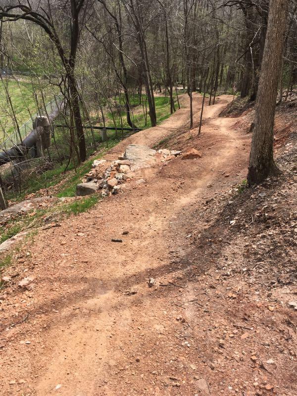 A winding dirt path through a forested area, surrounded by tall trees and patches of grass. The trail has scattered rocks and a slight incline, leading uphill. A grassy field and a pipe can be seen in the background. The scene is set in early spring, with bare tree branches hinting at new growth. Slaughter Pen Trail mountain bike trail.