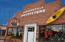 A brick storefront with the sign "GUNTERSVILLE OUTFITTERS" prominently displayed above. The front features large windows showcasing outdoor apparel and gear. A bright yellow kayak is mounted on the side of the building.
