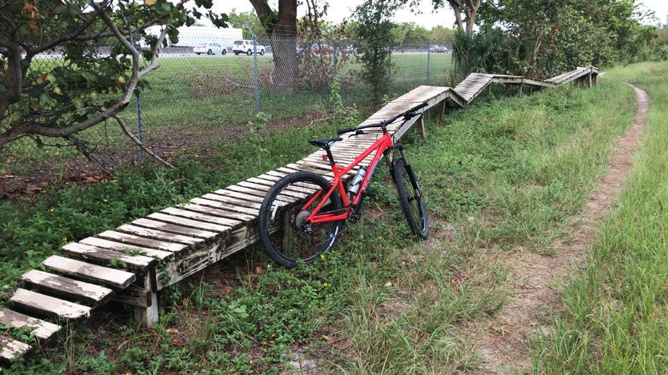 A red mountain bike resting on a wooden ramp surrounded by grassy terrain and trees, with a fence and vehicles visible in the background. The ramp has a zigzag design, indicating a biking trail. Quiet Waters Park mountain bike trail.
