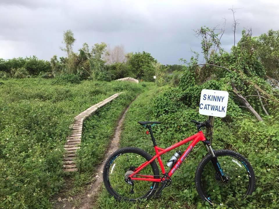 A red mountain bike is leaning against a sign that reads "Skinny Catwalk." The bike is positioned next to a narrow wooden path that winds through a grassy area with lush vegetation, while dark clouds loom in the sky above. Quiet Waters Park mountain bike trail.