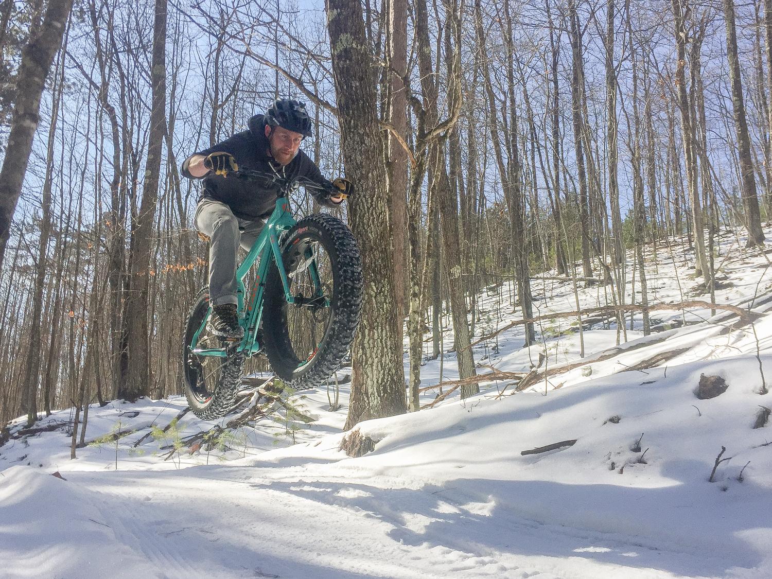 A person performing a jump on a fat bike in a snowy forest. The biker is wearing a helmet and gloves, with a focused expression, as they navigate through the winter landscape with tall, bare trees in the background and a snow-covered ground. LAMBO Zip Trails mountain bike trail.