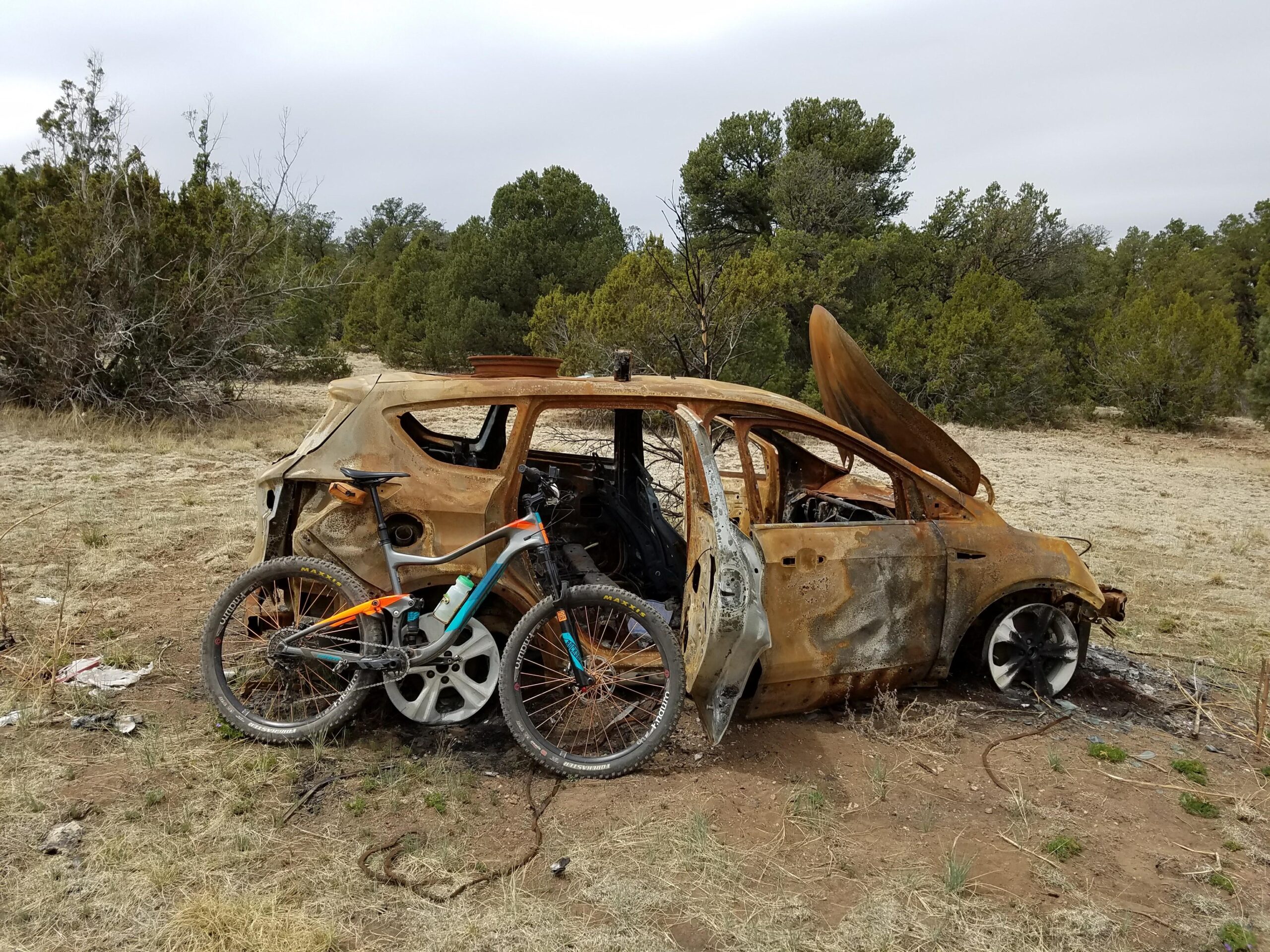 A rusty, abandoned car with an open hood sits in a grassy area surrounded by trees. A mountain bike is propped against the side of the car, which shows signs of damage and deterioration. The overcast sky adds to the desolate atmosphere of the scene.