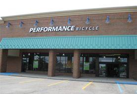Exterior view of a brick building with a green awning, featuring the sign "PERFORMANCE BICYCLE" above the entrance. The storefront has large glass windows and a parking lot in front.