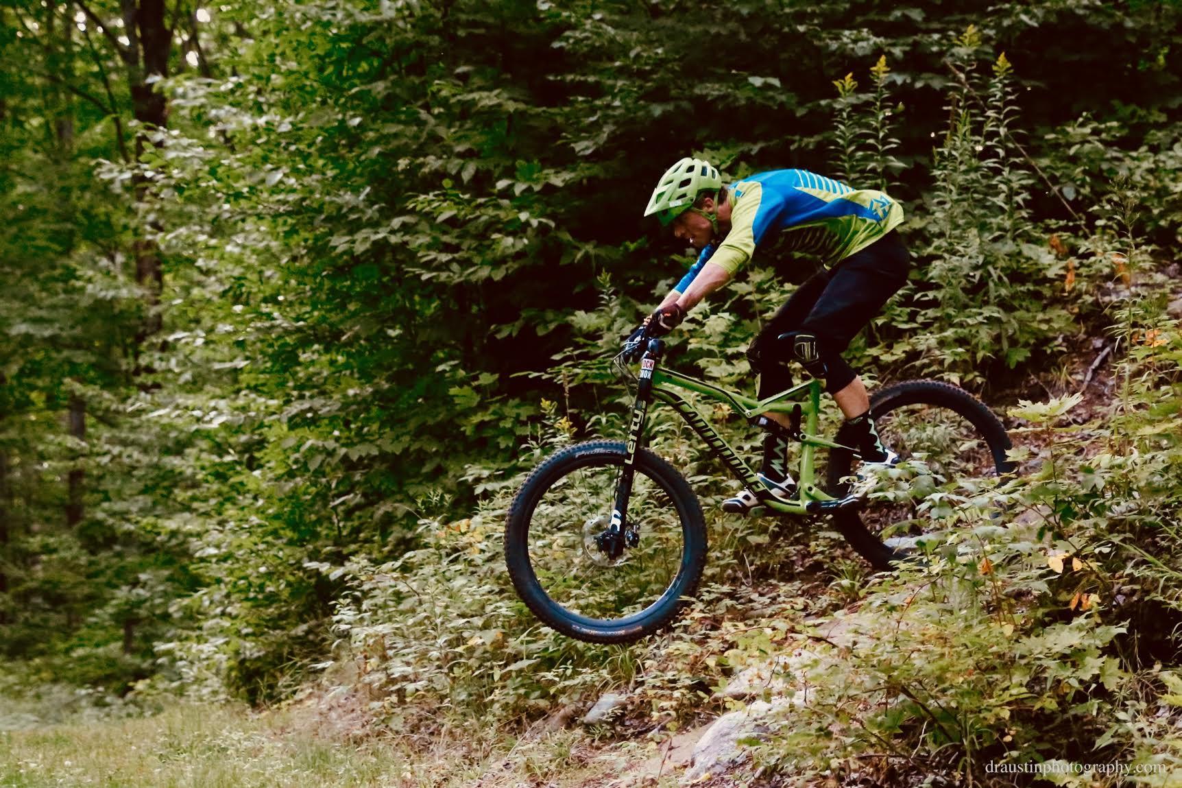 A mountain biker in a green and blue jersey and green helmet performs a jump on a bike trail surrounded by lush greenery. WinMan Trails mountain bike trail.