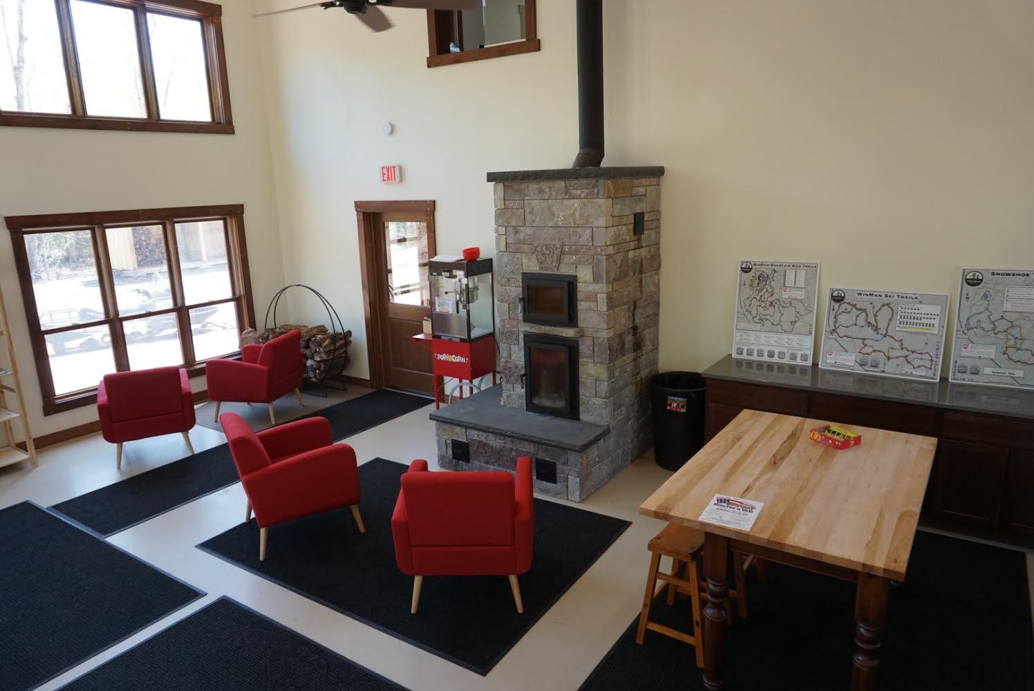 A cozy indoor seating area with red chairs arranged around a stone fireplace. Large windows allow natural light to fill the space, and there are maps displayed on a table against the wall. A wooden table with stools sits in the corner, completing the inviting atmosphere. WinMan Trail mountain bike trail.
