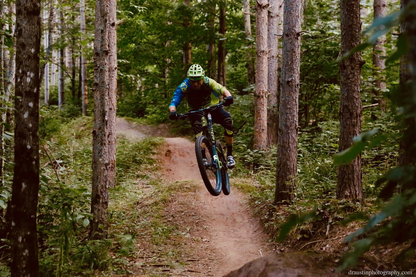 A mountain biker wearing a blue and green jersey jumps off a dirt ramp, surrounded by tall trees and lush greenery on a forest trail. WinMan Trails mountain bike trail.
