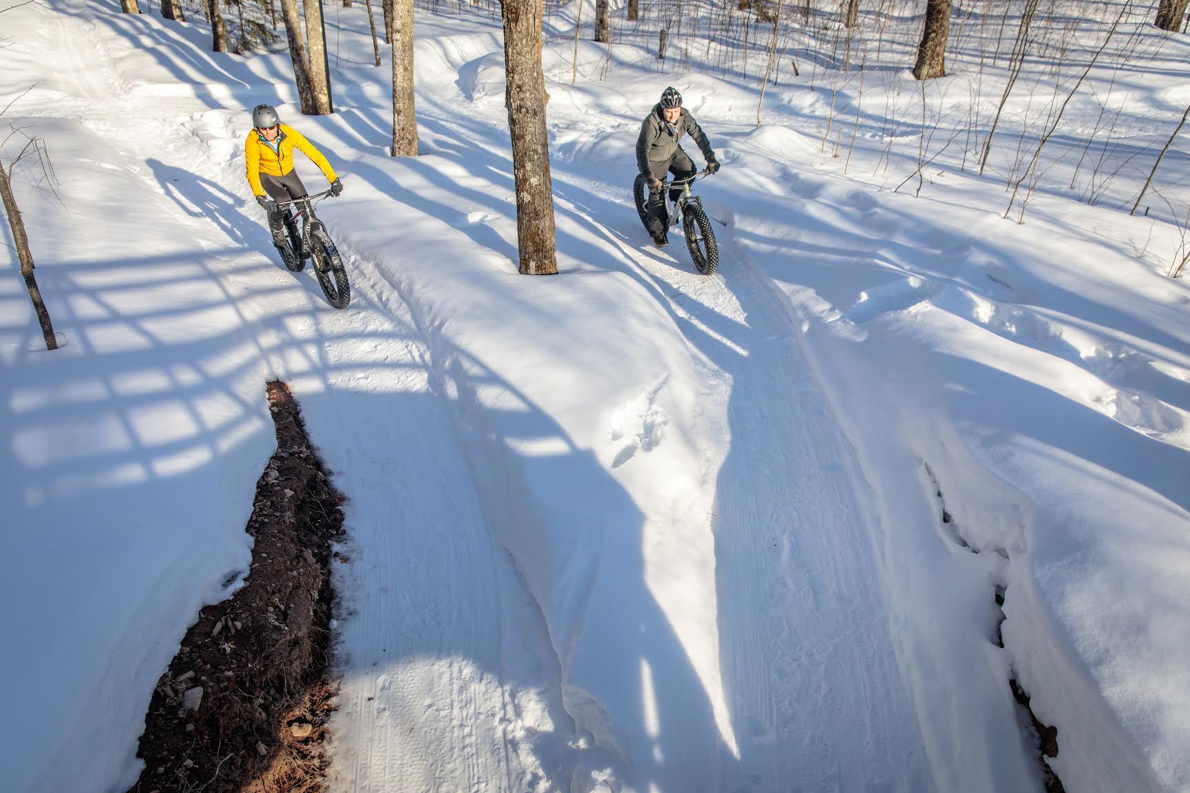 Two cyclists ride fat-tire bikes on a snowy trail through a forest. One cyclist, dressed in a yellow jacket, is navigating a track on the left, while the other, in a grey jacket, rides on the right. The scene is set amidst tall trees, with snow-covered ground and shadows creating a picturesque winter landscape. WinMan Trails mountain bike trail.