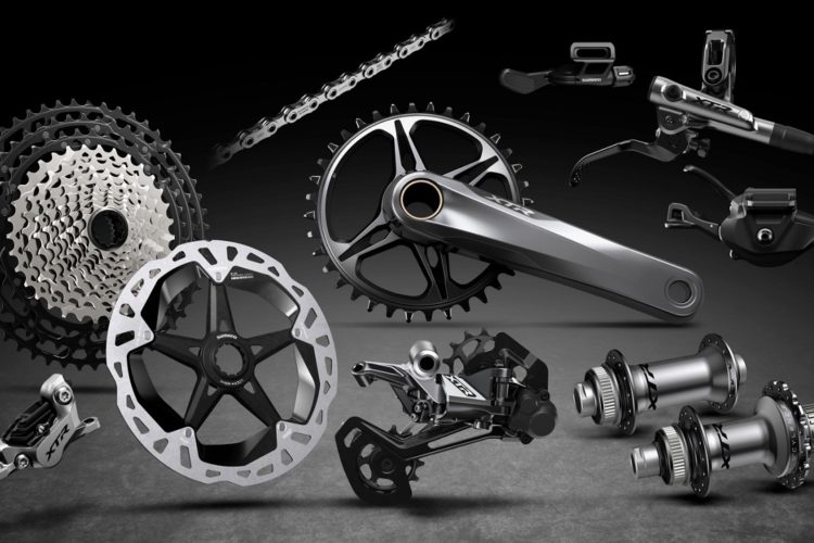 Various bicycle components arranged artistically, including a chain, cassette, crankset, disc brake rotors, derailleur, and hubs, all set against a dark background. The parts display sleek designs and are representative of high-performance cycling gear.