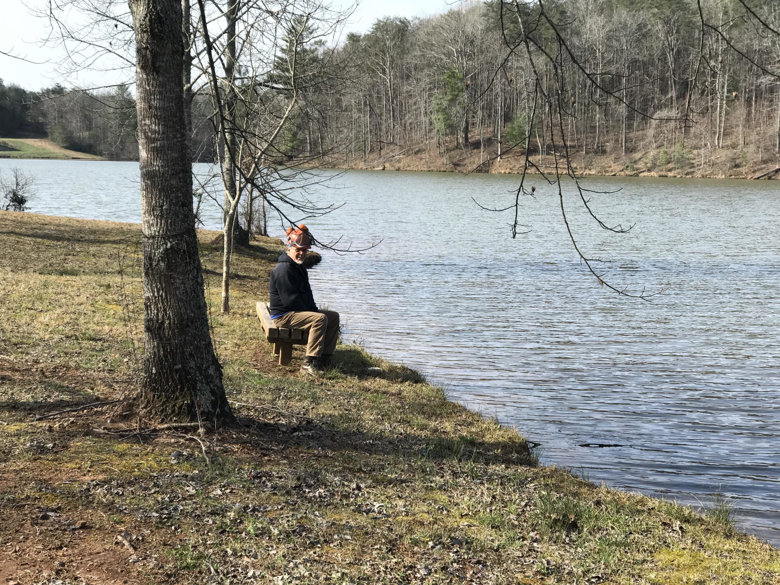 A person sitting on a wooden bench by the edge of a calm lake, surrounded by trees and a grassy bank. The individual is wearing a black jacket and has a hat on, looking toward the water. The background features a wooded area, partially reflecting in the lake