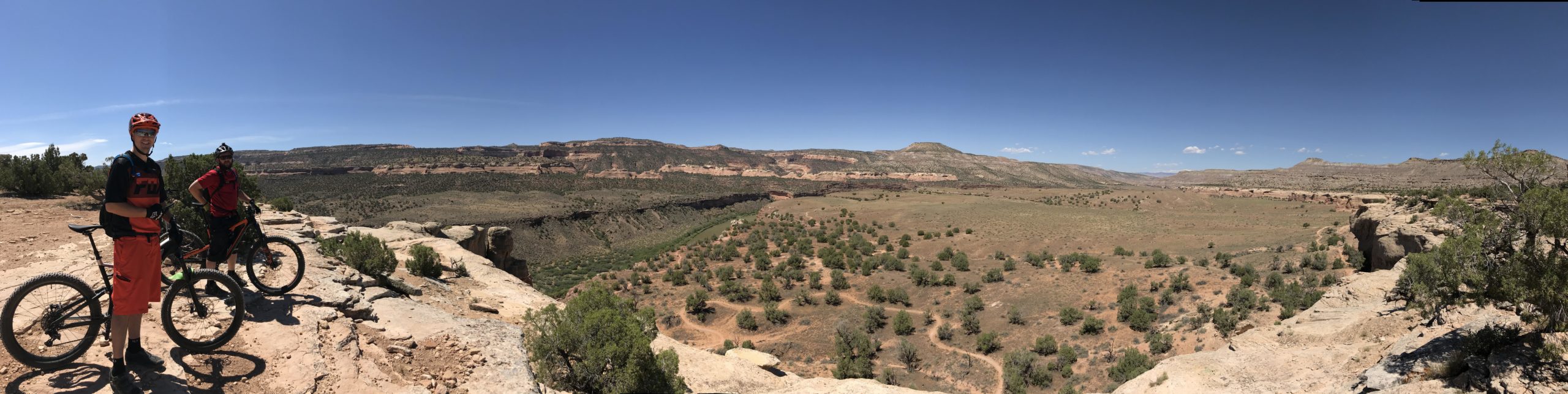 Two mountain bikers pause on a rocky ledge, overlooking a vast landscape of hills and valleys under a clear blue sky. The terrain features sparse vegetation, including small shrubs and scattered trees, set against a backdrop of cliffs and distant mountains. One biker is wearing an orange helmet and gear, while the other is dressed in red. The image captures the essence of outdoor adventure and the beauty of nature. Kokopelli Area Trails mountain bike trail.