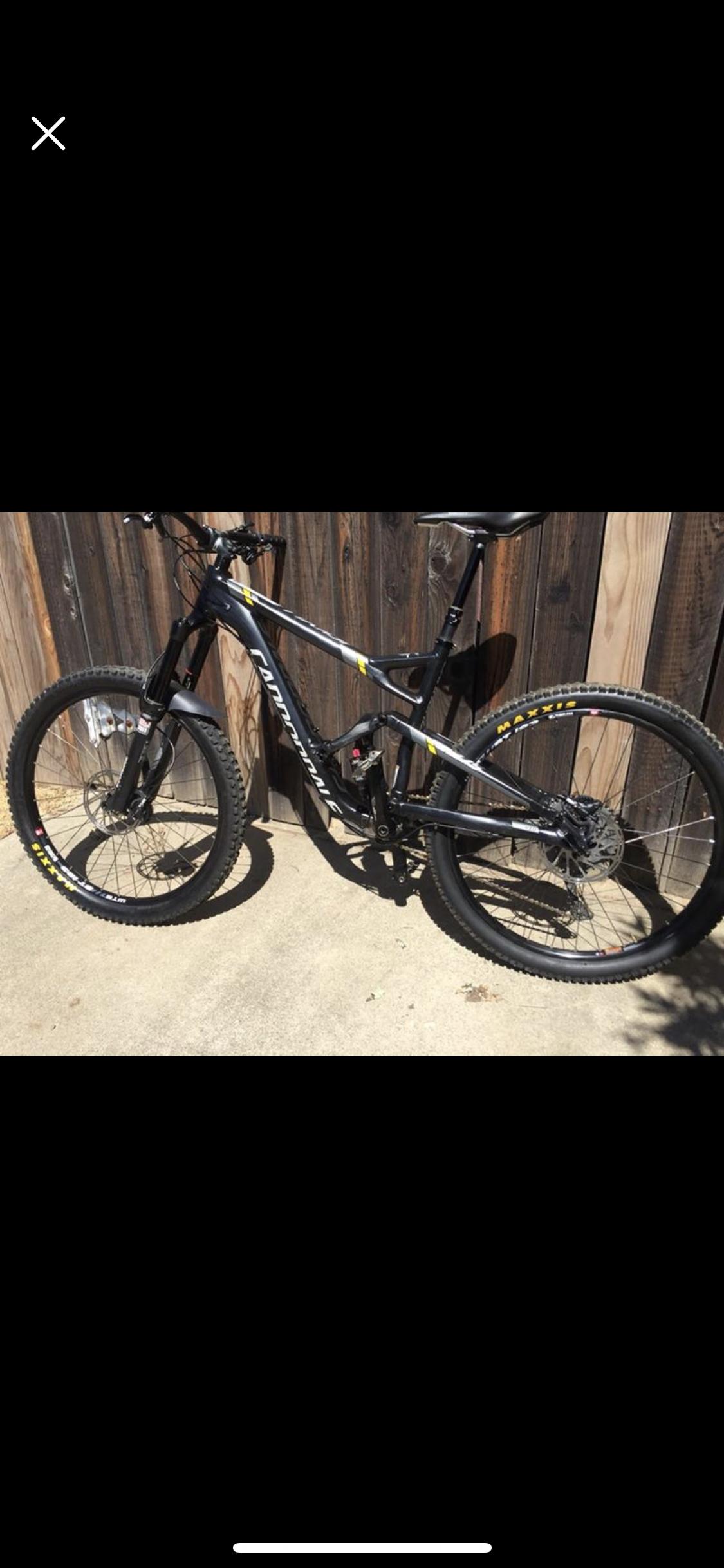 Cannondale Jekyll 3: A black mountain bike leaning against a wooden fence, showcasing its sturdy frame and thick, knobby tires. The bike features a sleek design with yellow accents and appears well-maintained, ready for outdoor adventures. The ground beneath it is paved, indicating a suburban setting.