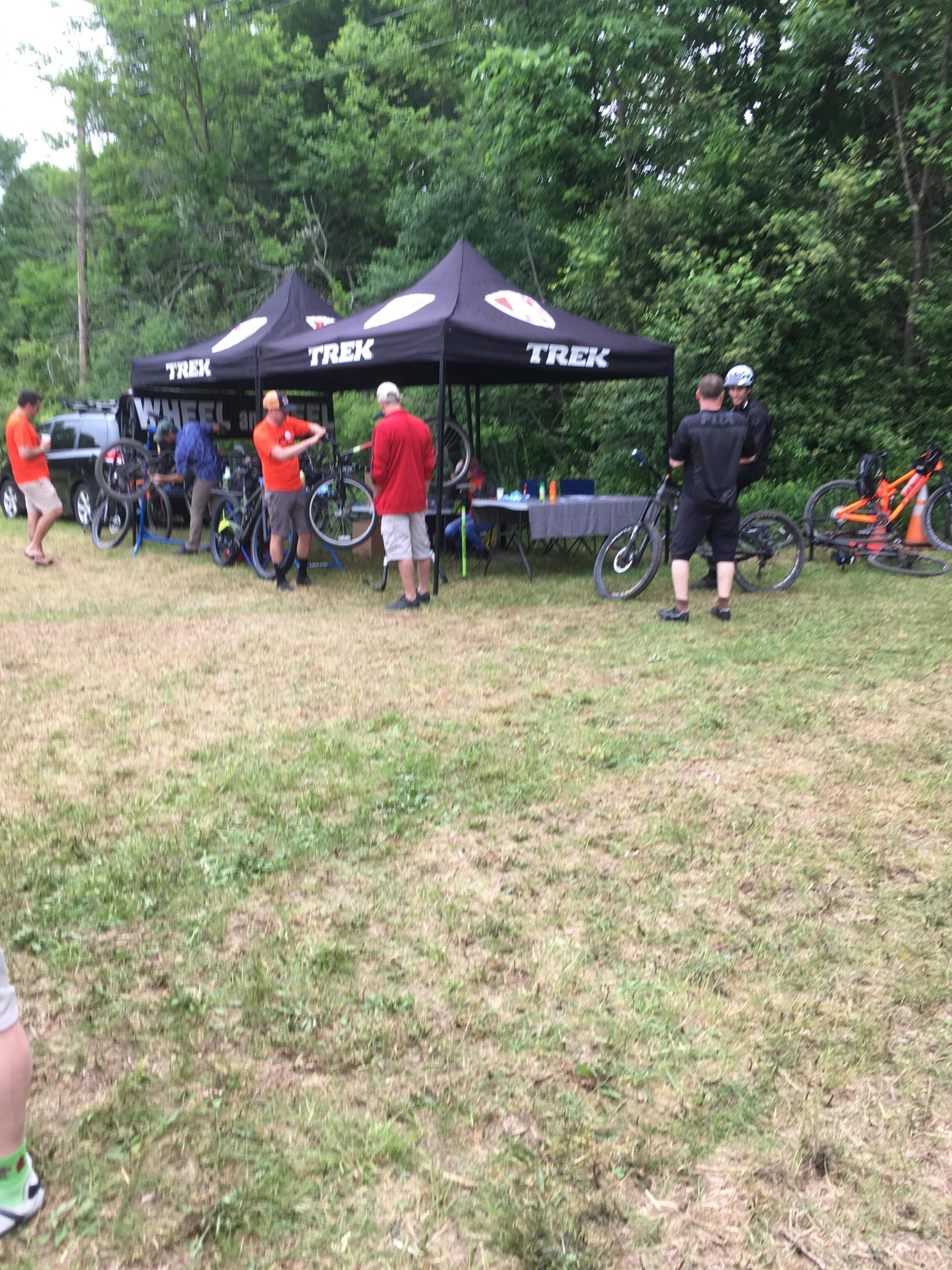 A group of people interacting around a Trek bicycle tent set up in a grassy area with trees in the background. Some individuals are adjusting bicycles, while others are engaged in conversation. The tent displays the Trek logo, and various bicycles are positioned nearby. Stewart State Forest mountain bike trail.