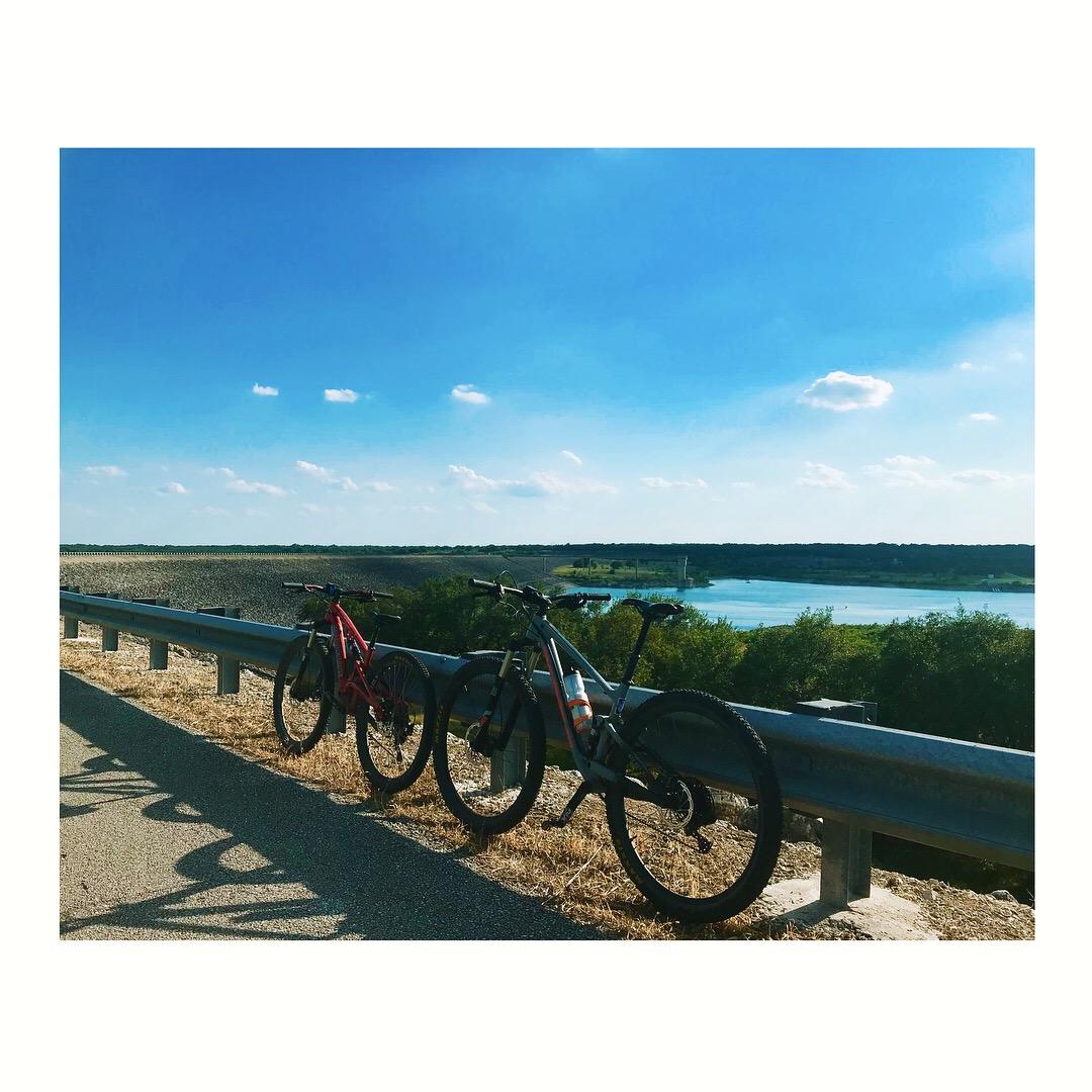 Two bicycles, one red and one gray, lean against a guardrail overlooking a scenic view of a lake and lush greenery under a bright blue sky with scattered clouds. The sun casts gentle shadows on the pavement, enhancing the peaceful outdoor setting. Goodwater Trail mountain bike trail.