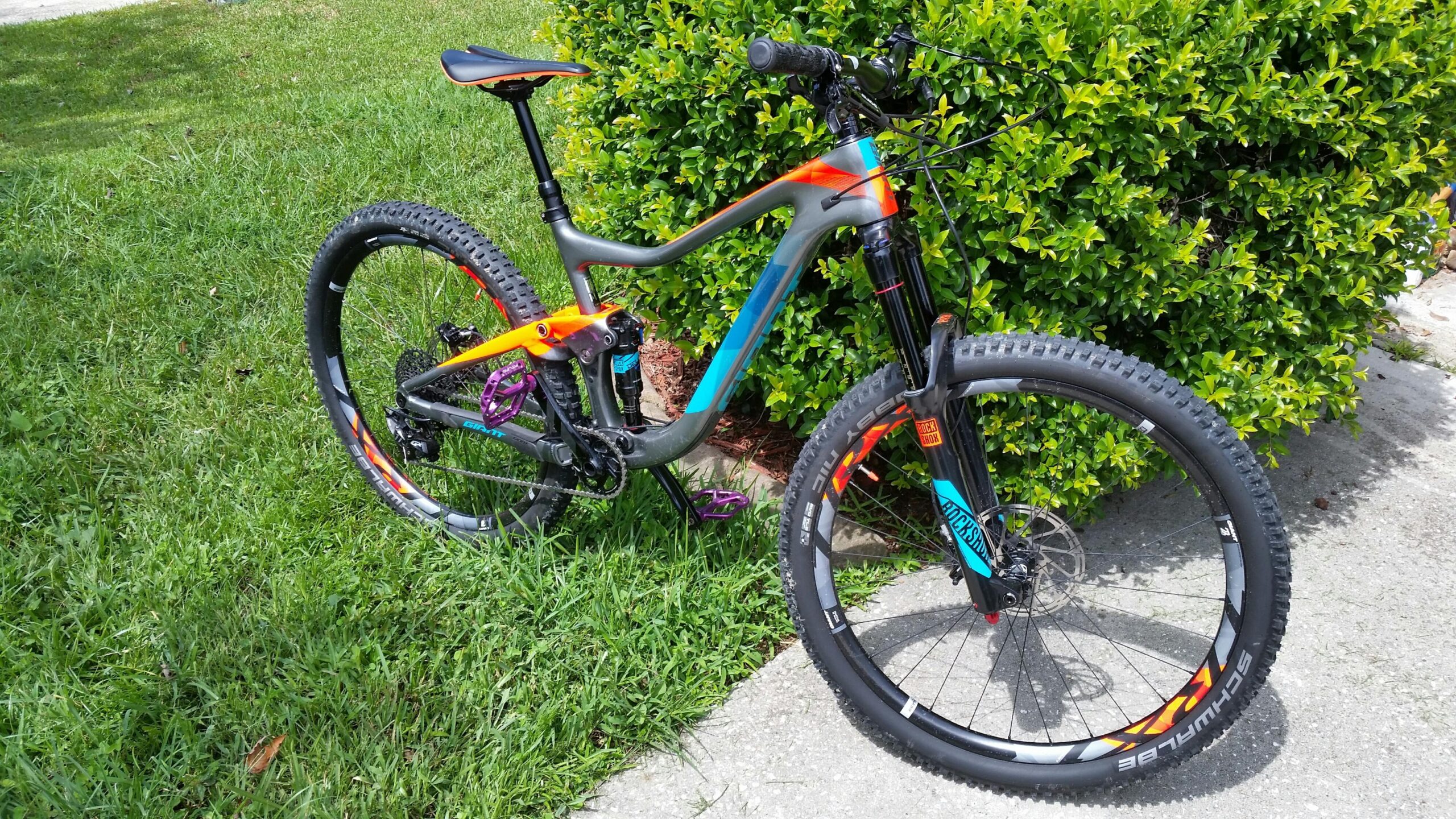 Giant Giant Trance Advanced 27.5 2: A colorful mountain bike with a gray frame featuring orange and blue accents, positioned on grass next to a bush. The bike has thick, knobby tires and a suspension fork, ideal for off-road riding.