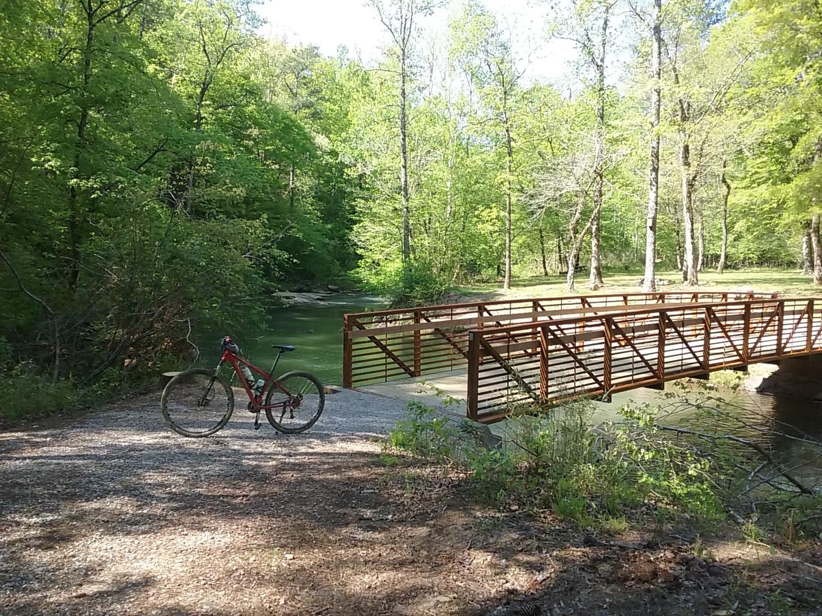 A red mountain bike is parked on a gravel pathway next to a calm green river, with a wooden bridge crossing over it. Surrounding the scene are lush green trees and bright sunlight filtering through the leaves, creating a serene outdoor atmosphere. Tannehill Historic Ironworks State Park mountain bike trail.