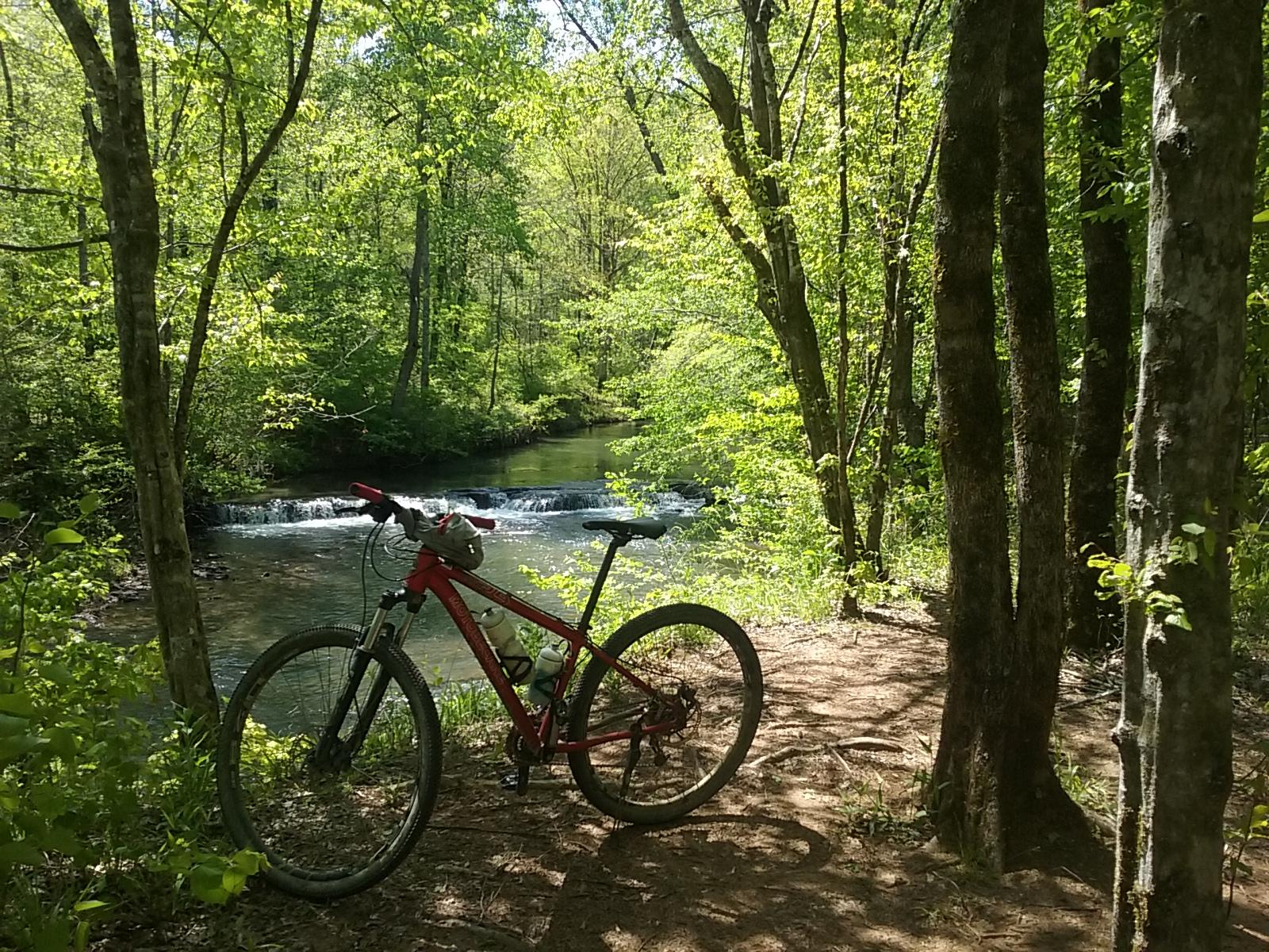 A red mountain bike resting on a dirt path beside a clear stream, surrounded by lush green trees and foliage on a sunny day. In the background, a small waterfall is visible. Tannehill Historic Ironworks State Park mountain bike trail.