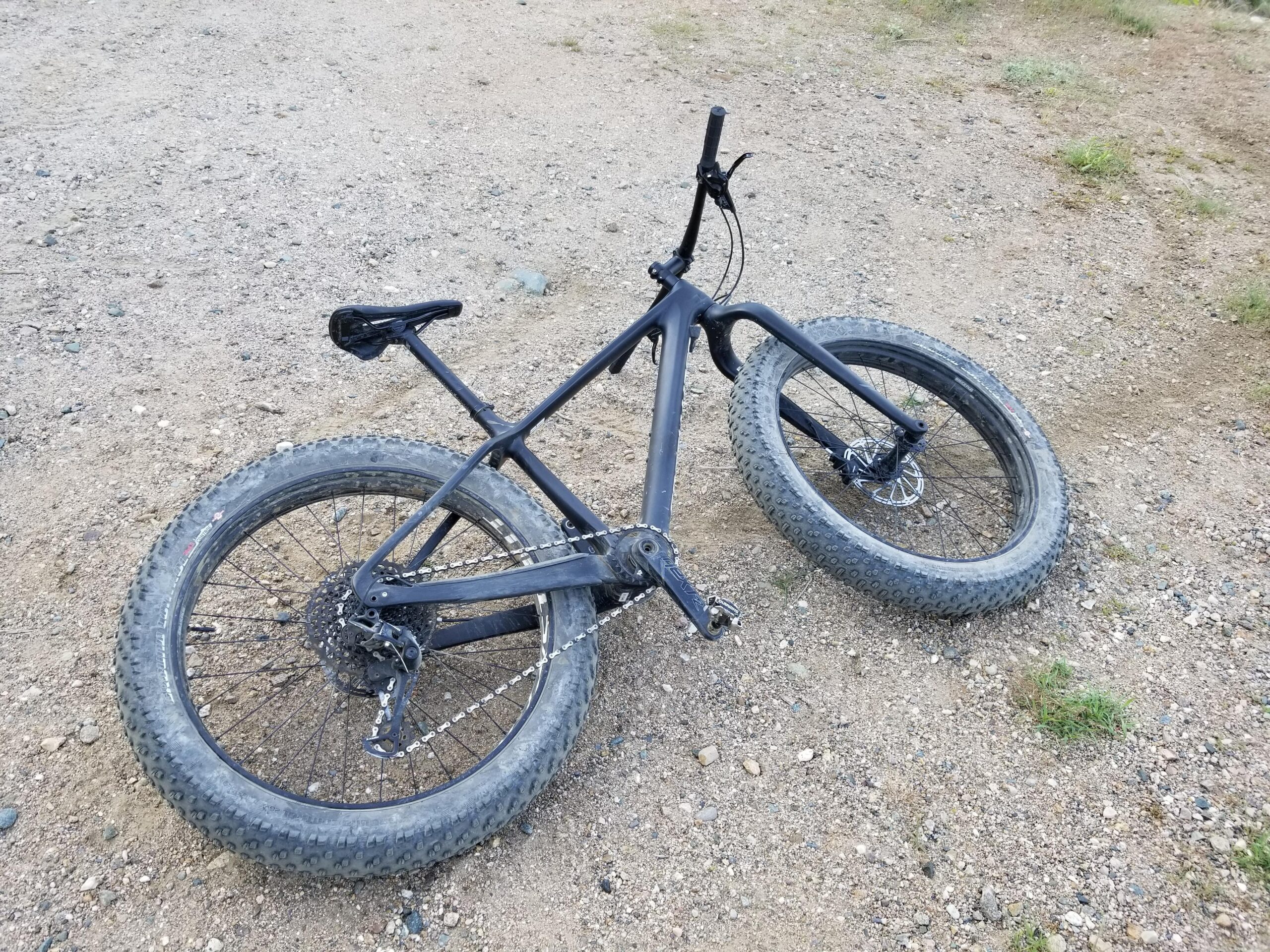 Specialized Fatboy Carbon Comp: A black fat tire bike laying on a gravel path, with its front wheel oriented to the right and the back wheel slightly off to the left. The terrain is sandy with some small pebbles and patches of grass.