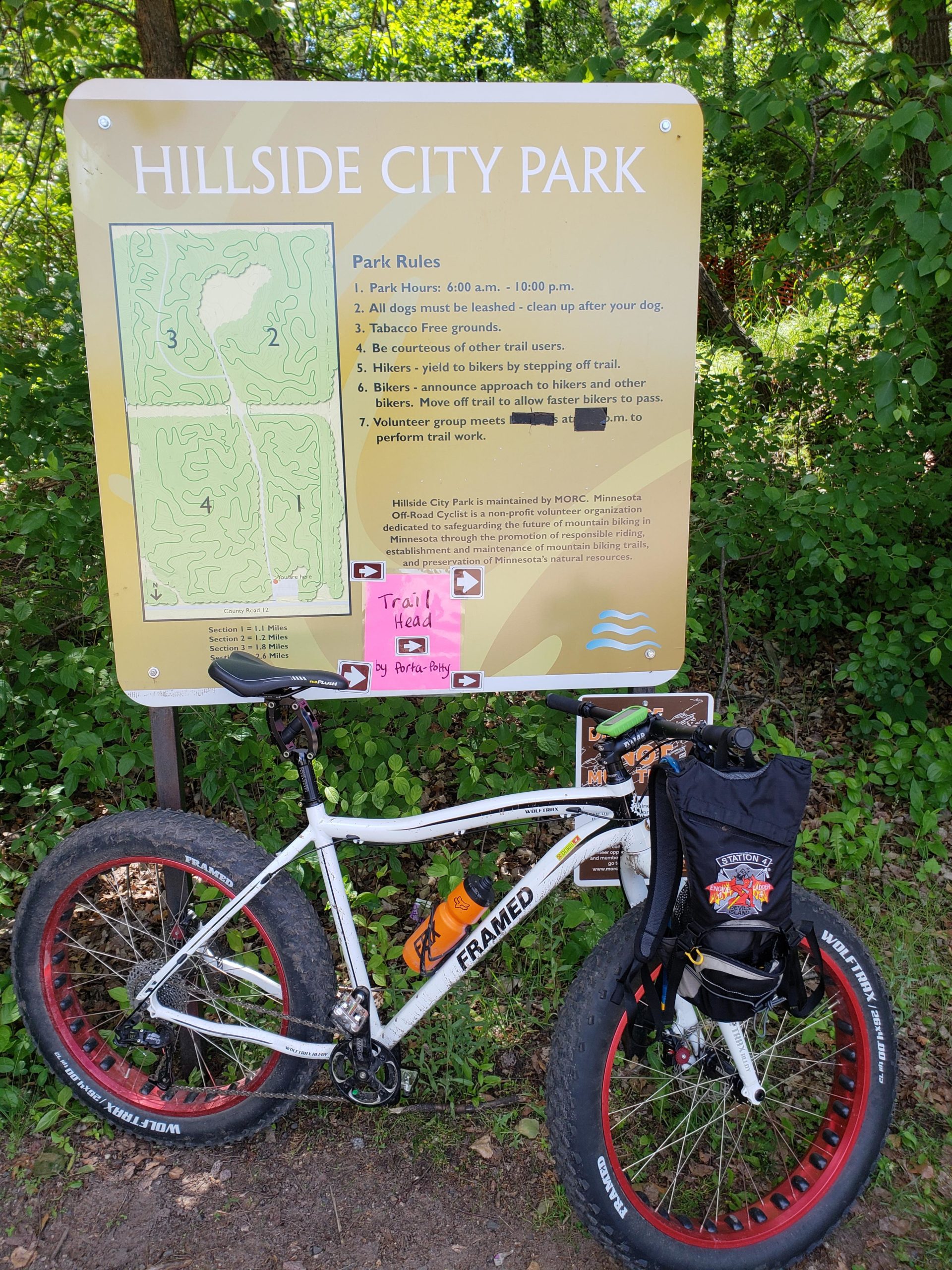 A white fat-tire bicycle parked next to a sign at Hillside City Park. The sign provides park rules, map details, and information about trail usage. Lush greenery surrounds the area, and a pink note indicating "Trail Head" is visible. Hillside Park mountain bike trail.