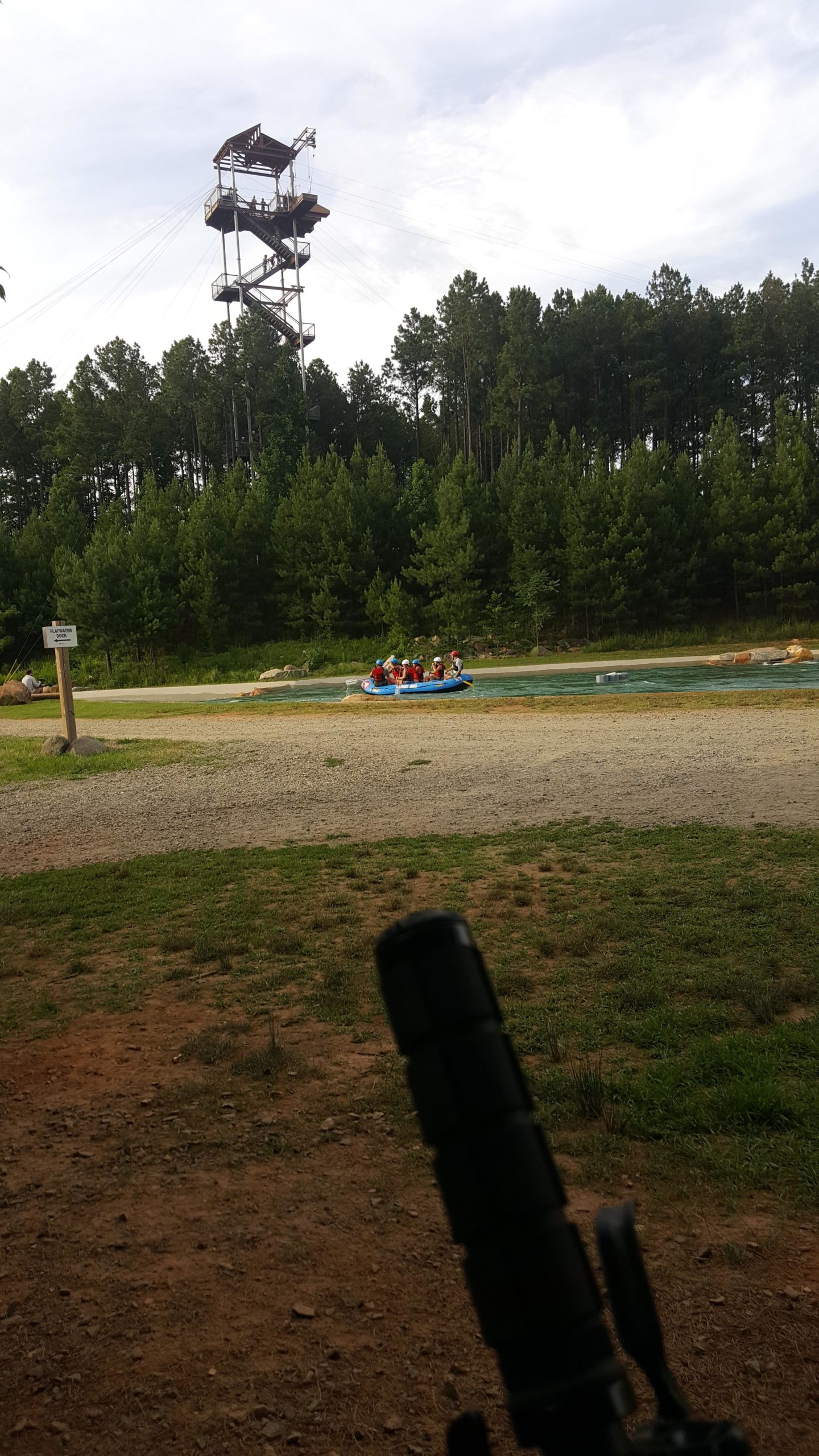 A group of people in a blue raft navigating a watercourse, with a tall climbing tower visible in the background surrounded by pine trees. The setting includes a gravel path and grassy areas near the water's edge. USNWC mountain bike trail.
