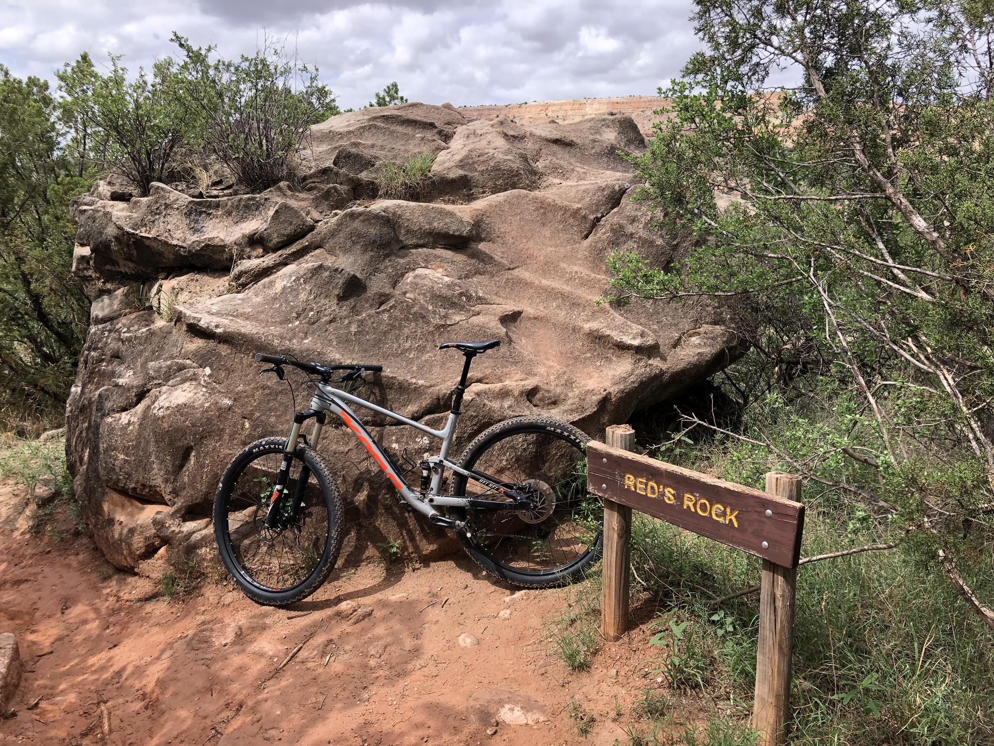 A mountain bike leaning against a large rock formation next to a wooden sign that reads "RED'S ROCK." The scene is surrounded by greenery, with a dirt path and a cloudy sky in the background. Palo Duro Canyon mountain bike trail.