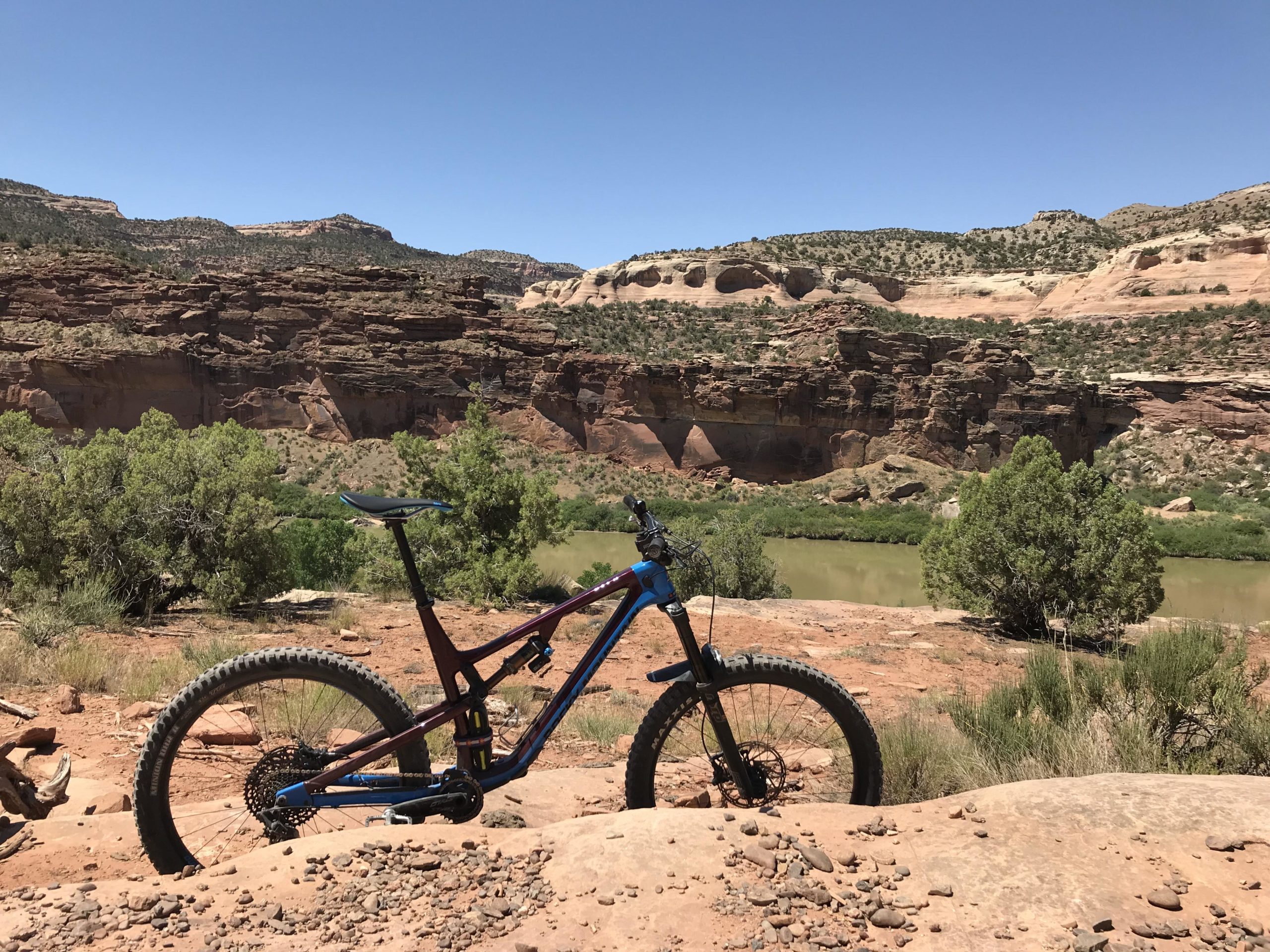 Mountain bike resting on rocky terrain with a scenic backdrop of red rock formations and lush greenery along a river. The clear blue sky adds to the vibrant outdoor setting. Kokopelli Area Trails mountain bike trail.